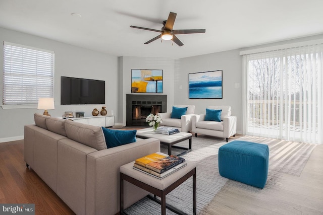 living area featuring a ceiling fan, dark wood-type flooring, a fireplace with raised hearth, and baseboards