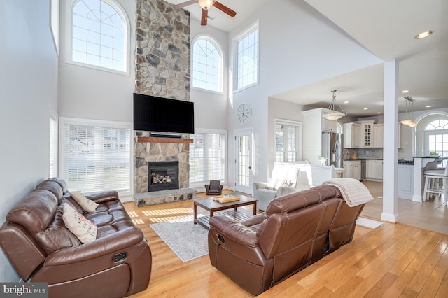 living area featuring a stone fireplace, light wood-style flooring, a high ceiling, and a ceiling fan