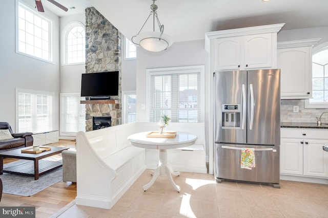 kitchen with stainless steel refrigerator with ice dispenser, backsplash, dark countertops, white cabinetry, and a stone fireplace