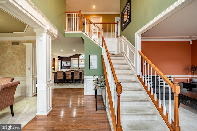 stairway with ornamental molding, wood finished floors, ornate columns, and wainscoting