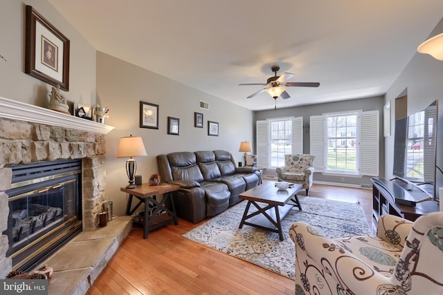 living room with hardwood / wood-style floors, a ceiling fan, visible vents, baseboards, and a stone fireplace