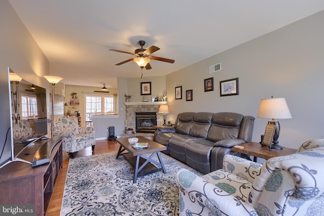 living room with visible vents, wood finished floors, a stone fireplace, baseboards, and ceiling fan