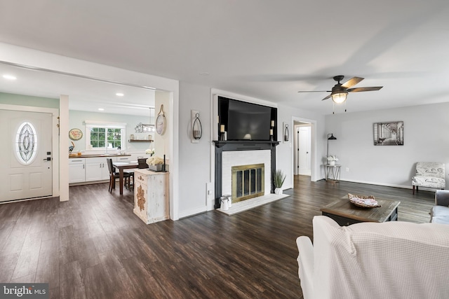 living area featuring baseboards, recessed lighting, dark wood-style flooring, ceiling fan, and a brick fireplace