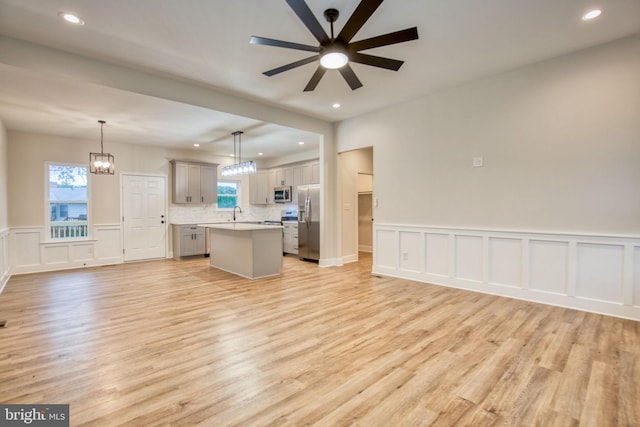 unfurnished living room with light wood finished floors, a wainscoted wall, recessed lighting, ceiling fan with notable chandelier, and a decorative wall