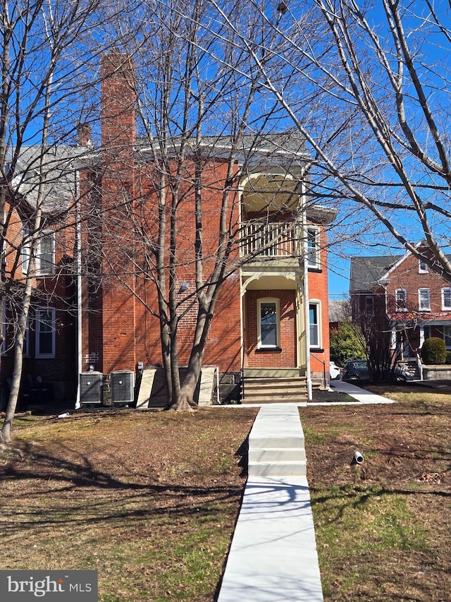 view of front of property featuring a balcony, central AC unit, brick siding, and a chimney