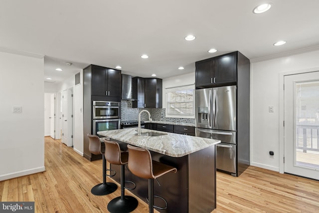 kitchen with light stone countertops, decorative backsplash, light wood-style floors, stainless steel appliances, and wall chimney exhaust hood