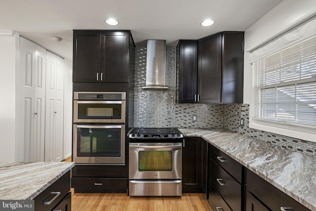 kitchen with light stone counters, stainless steel appliances, light wood-style flooring, and wall chimney range hood