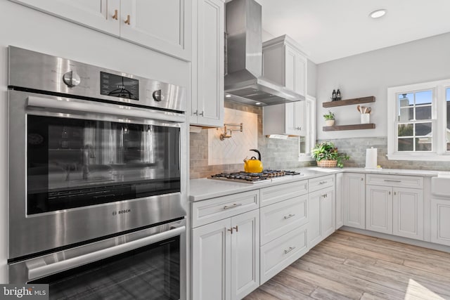 kitchen with light wood finished floors, light countertops, appliances with stainless steel finishes, wall chimney range hood, and tasteful backsplash