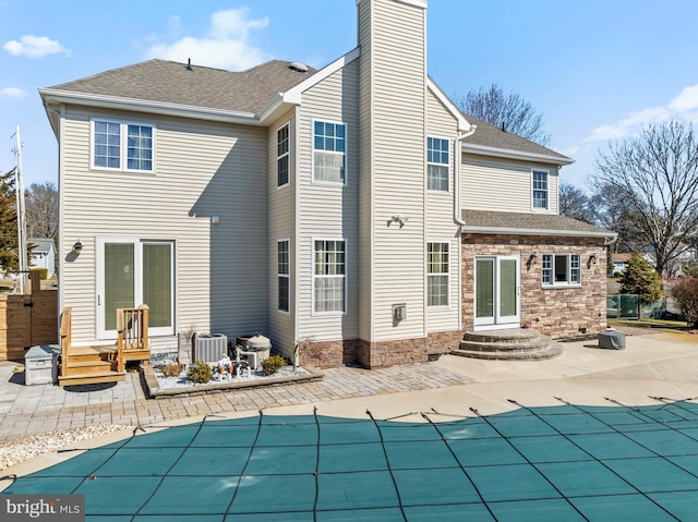 rear view of property with a patio, central AC, a shingled roof, a chimney, and stone siding