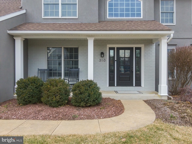 view of exterior entry featuring brick siding, covered porch, and roof with shingles