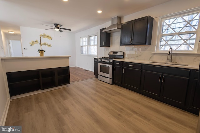 kitchen with light wood finished floors, stainless steel range with gas stovetop, a sink, wall chimney exhaust hood, and backsplash