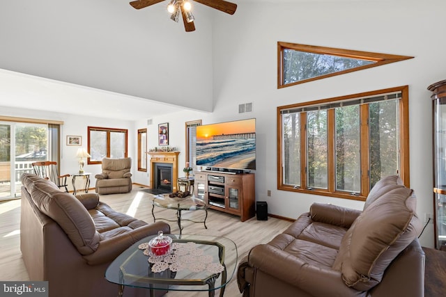 living room with a ceiling fan, visible vents, a fireplace with flush hearth, light wood-style floors, and a towering ceiling