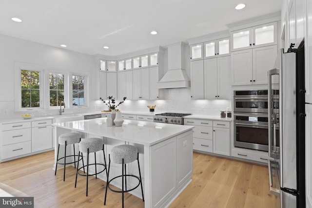 kitchen with premium range hood, a kitchen island, backsplash, appliances with stainless steel finishes, and light wood finished floors