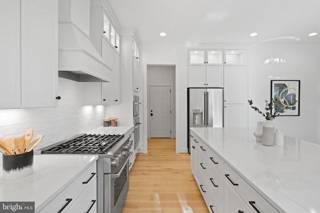 kitchen with premium range hood, high quality appliances, backsplash, white cabinetry, and light wood finished floors