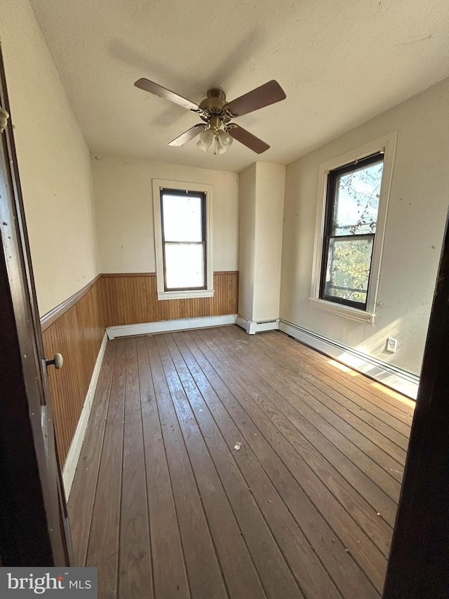 empty room featuring a ceiling fan, hardwood / wood-style flooring, wood walls, wainscoting, and a baseboard heating unit