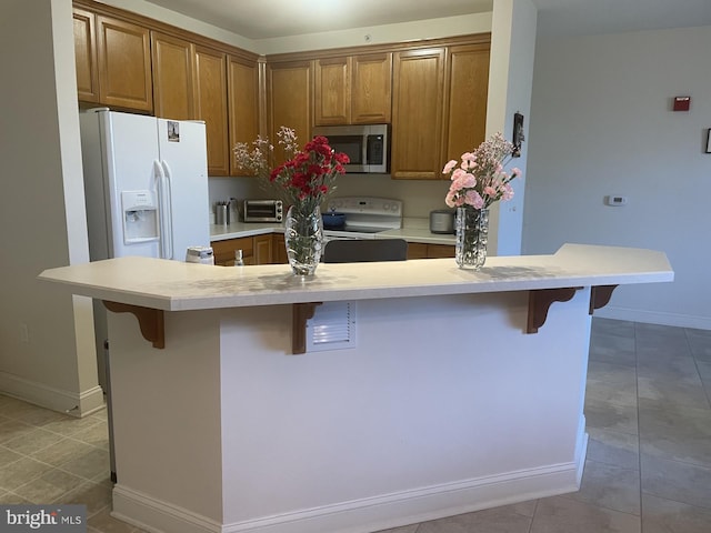 kitchen featuring a breakfast bar area, light countertops, light tile patterned floors, brown cabinets, and white appliances