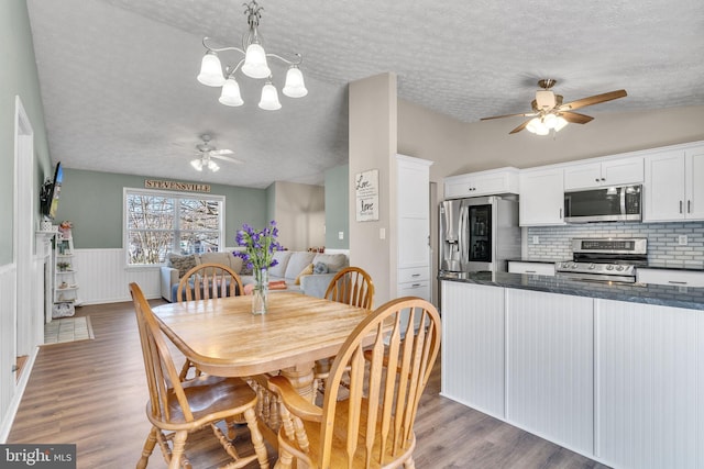 dining space with wood finished floors, ceiling fan with notable chandelier, a wainscoted wall, and a textured ceiling