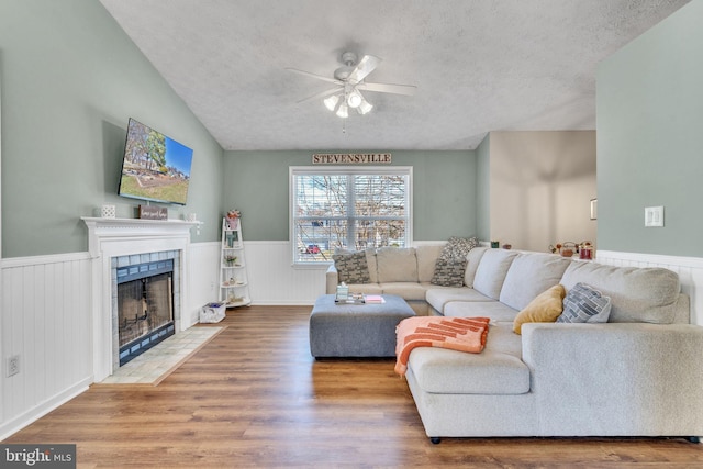 living room with wood finished floors, a wainscoted wall, ceiling fan, a tile fireplace, and a textured ceiling