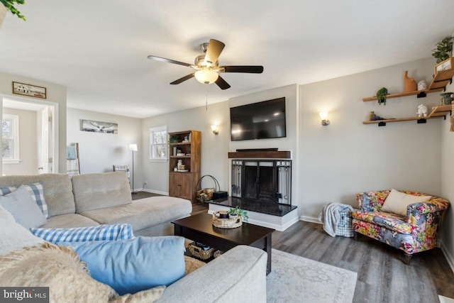 living area featuring dark wood-type flooring, a fireplace with raised hearth, baseboards, and ceiling fan