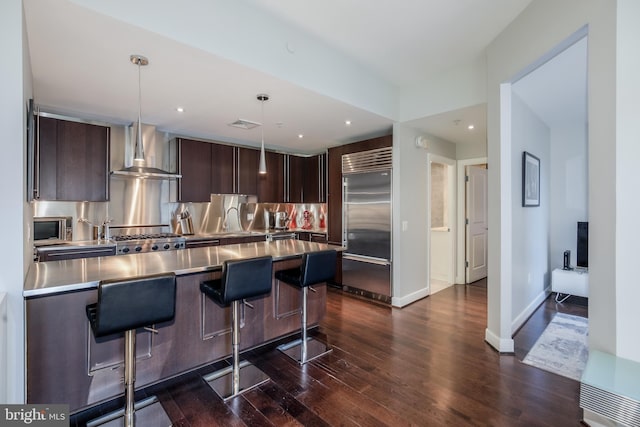 kitchen with stainless steel countertops, dark wood-style floors, stainless steel appliances, a breakfast bar area, and wall chimney range hood