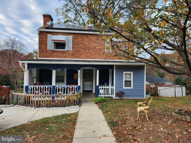 view of front of property with brick siding, fence, a porch, roof mounted solar panels, and a chimney