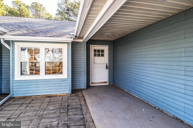 view of exterior entry featuring an attached carport, a shingled roof, and a patio