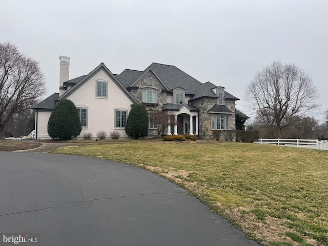 french country home featuring fence, a chimney, stucco siding, a front lawn, and stone siding