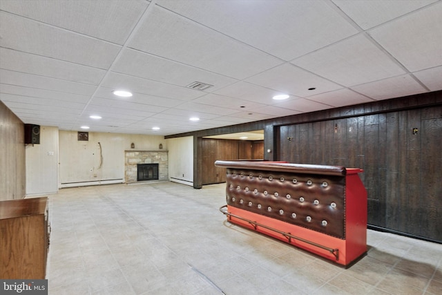 interior space with a drop ceiling, a baseboard radiator, a stone fireplace, and wood walls