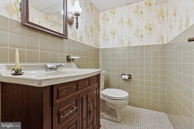 half bath with vanity, wallpapered walls, tile patterned flooring, tile walls, and toilet