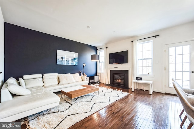 living room featuring a glass covered fireplace, baseboards, wood-type flooring, and an accent wall