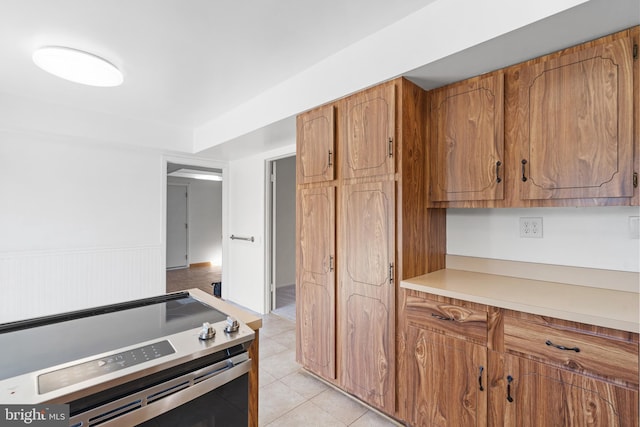 kitchen with brown cabinets, stainless steel electric range, light countertops, a wainscoted wall, and light tile patterned floors