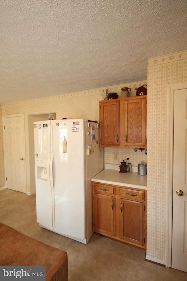 kitchen with wallpapered walls, light countertops, brown cabinets, white fridge with ice dispenser, and a textured ceiling