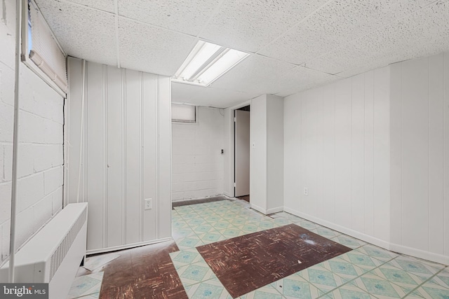 finished basement featuring tile patterned floors, a paneled ceiling, concrete block wall, and radiator heating unit