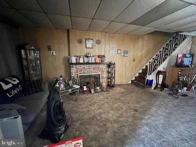 carpeted living area with a drop ceiling, stairway, a fireplace with flush hearth, and wooden walls