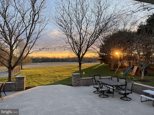 patio terrace at dusk featuring a playground, outdoor dining area, a lawn, and fence