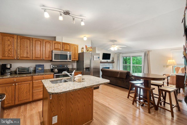 kitchen with a center island with sink, light stone counters, appliances with stainless steel finishes, brown cabinetry, and a sink