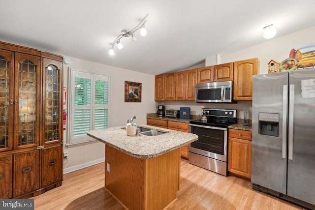 kitchen featuring light stone countertops, a center island with sink, light wood finished floors, a sink, and appliances with stainless steel finishes