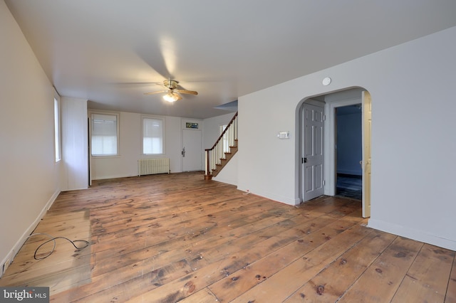 unfurnished living room featuring a ceiling fan, stairway, wood-type flooring, radiator, and arched walkways