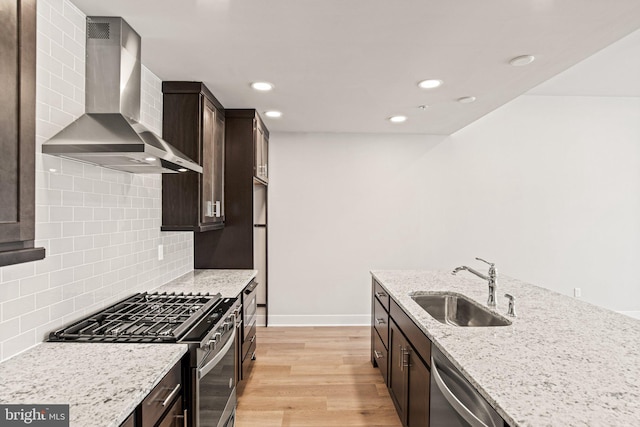 kitchen featuring wall chimney range hood, dark brown cabinetry, light wood-type flooring, appliances with stainless steel finishes, and a sink