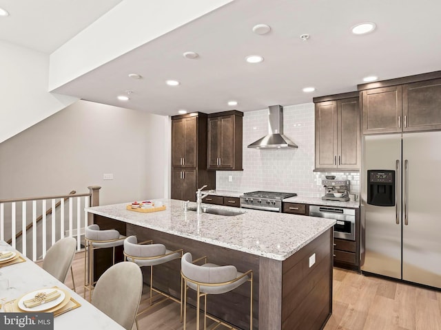 kitchen featuring light wood finished floors, dark brown cabinets, wall chimney range hood, stainless steel appliances, and a sink