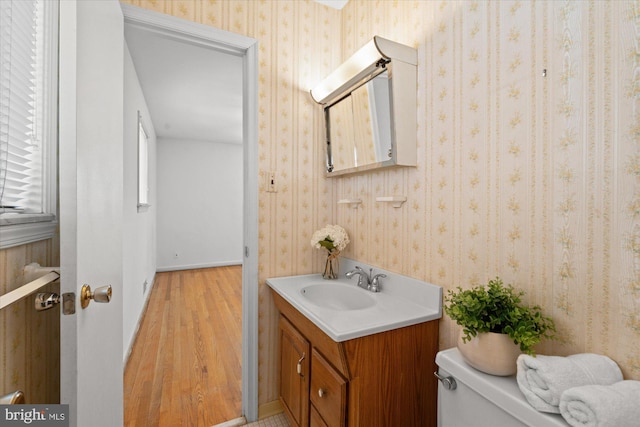 half bathroom with toilet, wood finished floors, vanity, and wallpapered walls