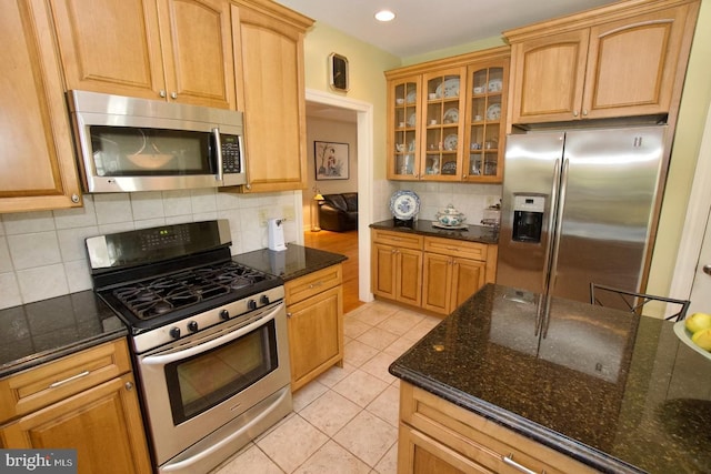 kitchen with dark stone counters, light tile patterned flooring, glass insert cabinets, appliances with stainless steel finishes, and tasteful backsplash