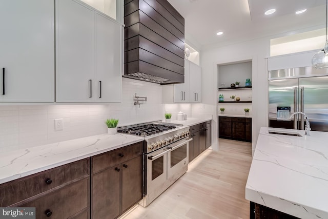 kitchen with custom range hood, a sink, premium appliances, open shelves, and light stone countertops