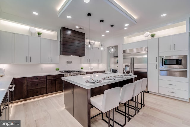 kitchen with a warming drawer, a breakfast bar, wall chimney exhaust hood, light wood finished floors, and built in appliances