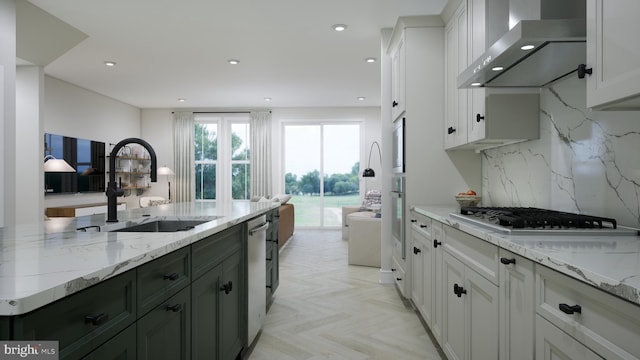 kitchen with wall chimney range hood, light stone countertops, dishwasher, white cabinets, and a sink