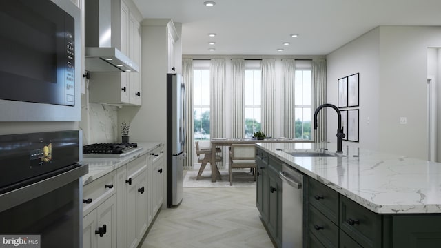 kitchen with a center island with sink, a sink, stainless steel appliances, white cabinets, and wall chimney exhaust hood