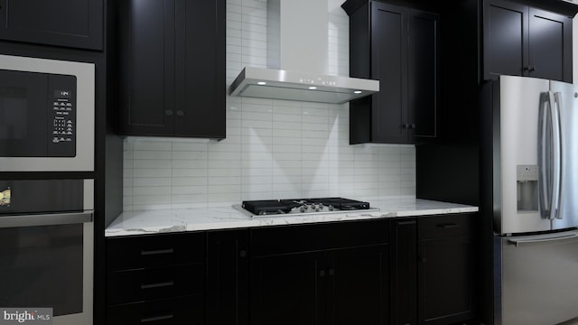 kitchen with backsplash, light stone counters, appliances with stainless steel finishes, dark cabinetry, and wall chimney exhaust hood