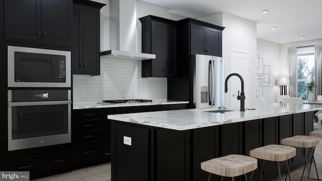 kitchen with wall chimney range hood, light stone countertops, decorative backsplash, stainless steel appliances, and a sink