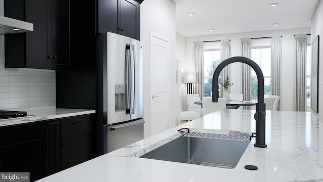 kitchen with tasteful backsplash, light stone countertops, stainless steel fridge with ice dispenser, range hood, and dark cabinets