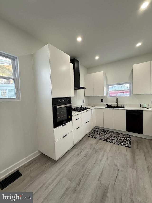 kitchen with visible vents, black appliances, a sink, wall chimney range hood, and light wood finished floors